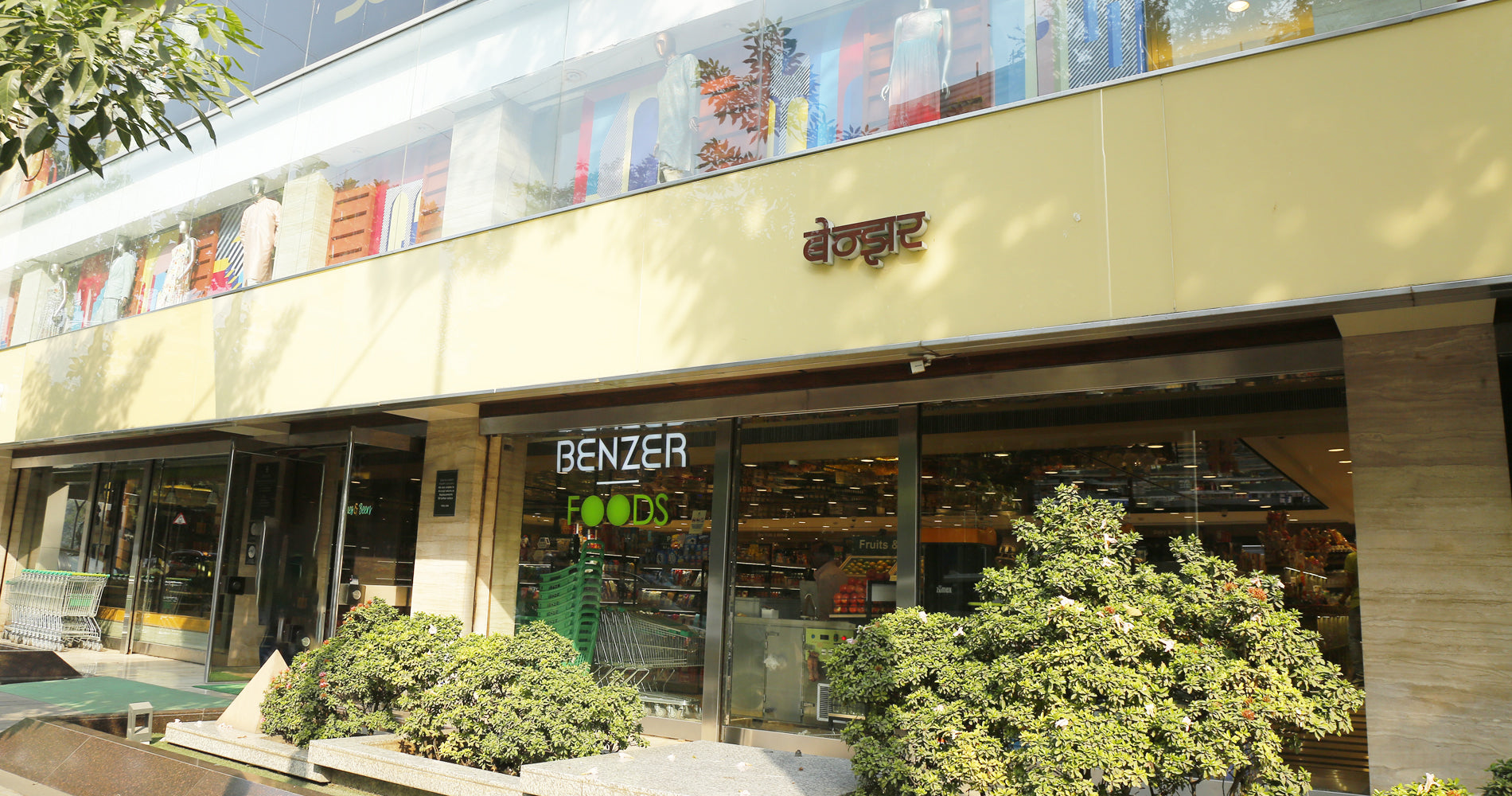A storefront facade with a beige upper section, large glass windows, and the visible brand name BENZER in white letters. Green shrubs line the front, and the interior of the store is visible through the glass.