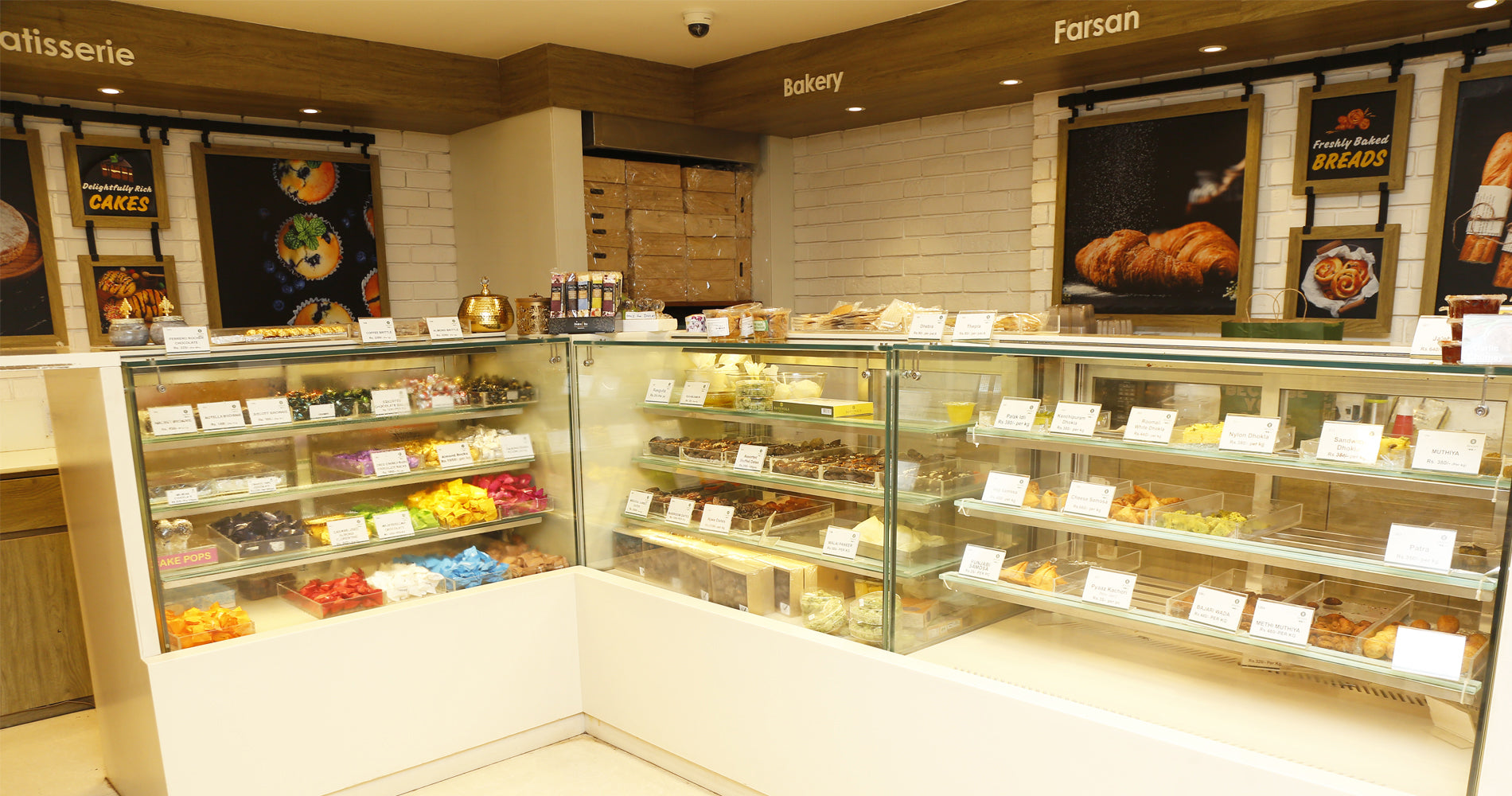 A bakery display case filled with assorted pastries, cakes, and sweets in a clean, modern pastry shop interior with white brick walls and wooden signage.