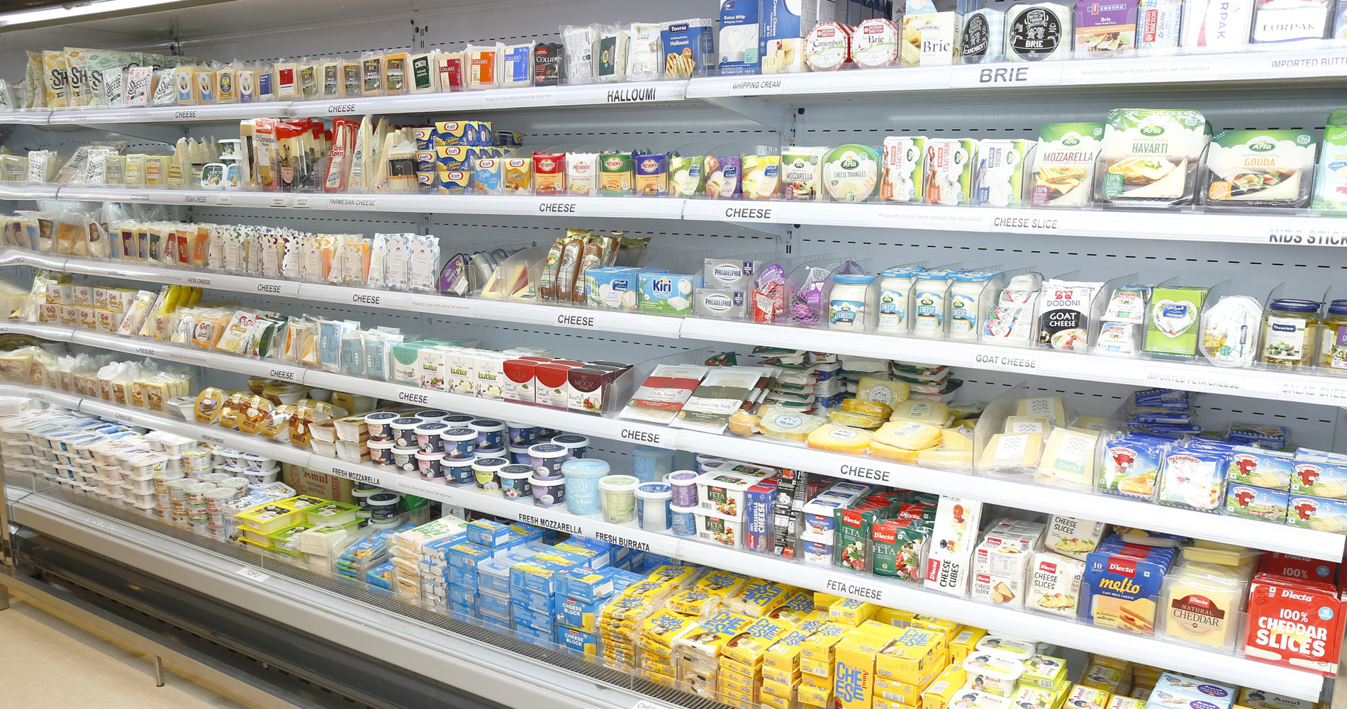 A well-stocked grocery shelf in a supermarket or deli, displaying various packaged foods including cheeses, crackers, yogurt, and baked goods.