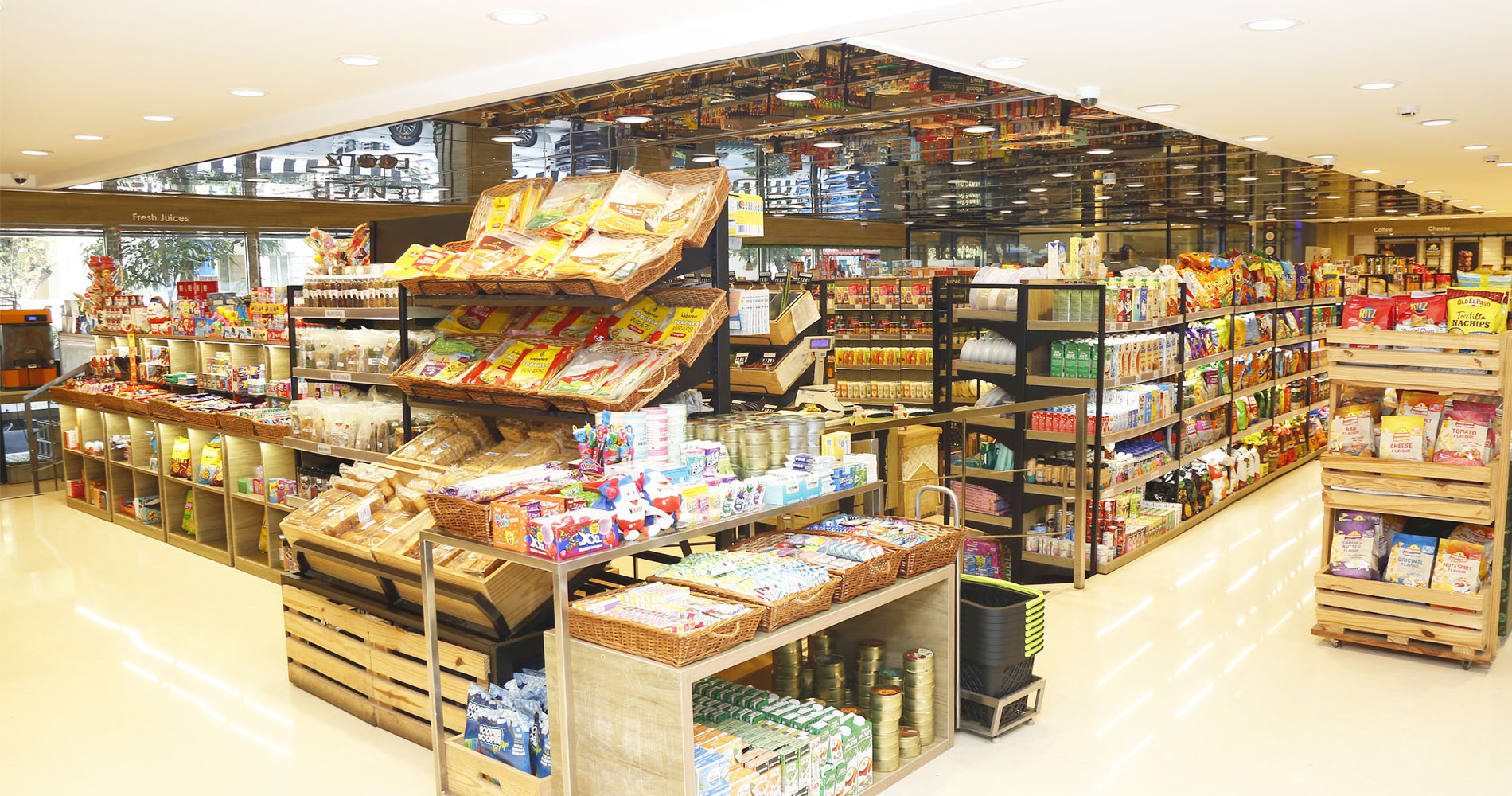A wide interior view of a well-lit grocery store aisle with wooden pallet display stands stocked with packaged snacks, breads, canned drinks, and other retail goods. Shelves on both sides are filled with assorted products under bright ceiling lights.