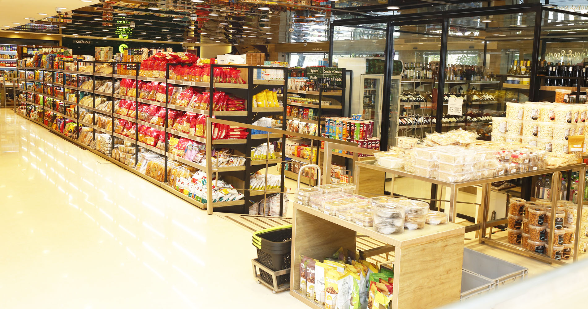 A wide interior view of a well-lit grocery store aisle with tall black shelving stocked with packaged snacks, breads, and other grocery items. Display tables in the foreground hold packaged goods and clear plastic containers, with a glass-front beverage or food section visible in the background.