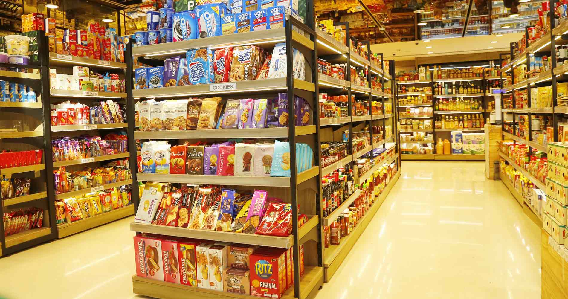 A brightly lit grocery store aisle with multiple shelves stocked with packaged snacks, cereals, and other retail products. The floor is glossy and reflects the overhead lighting, and no people are visible.