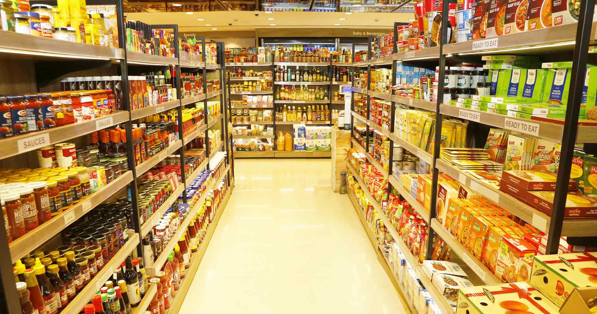 A brightly lit grocery store aisle with shelves stocked with packaged foods, sauces, and beverages on both sides.