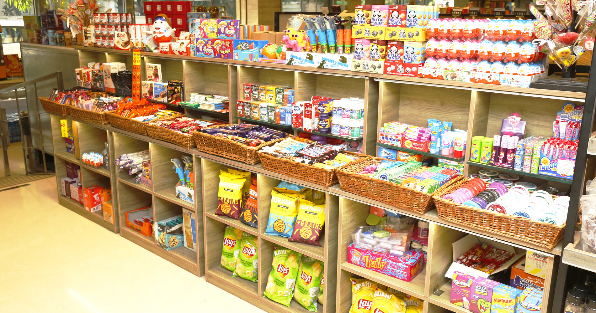 A retail candy and snack display shelf in a store, filled with packaged sweets, chips, and other confectionery items arranged on wooden cubbies and wicker baskets.